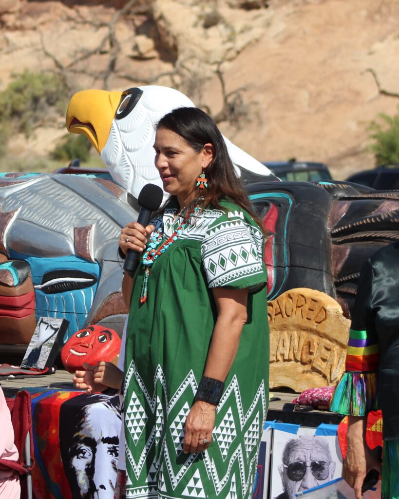Dr. Doreen Bird holds a microphone and talks to a crowd in her traditional dresswear.