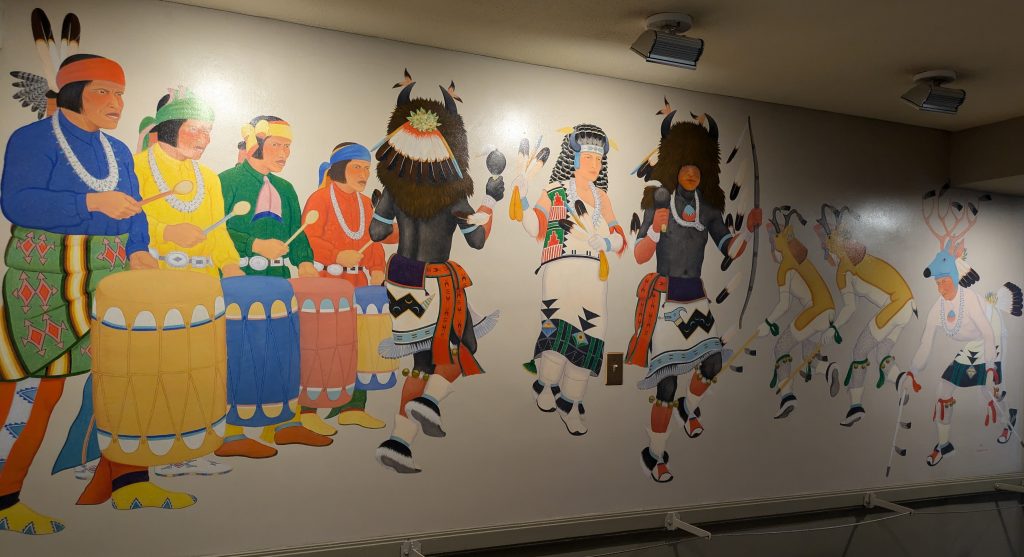 A painted mural of 4 Pueblo men drumming as 6 Pueblo men dance in traditional dance regalia.