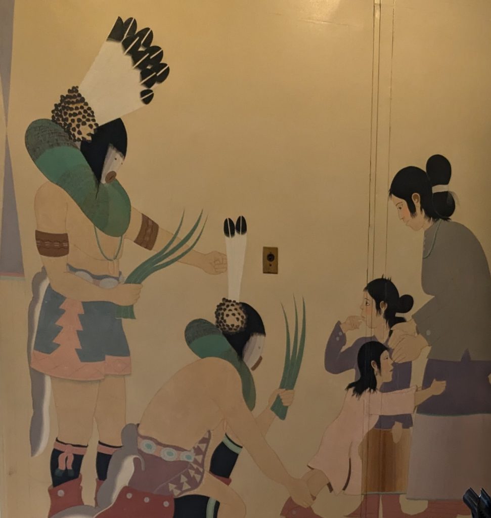 A painted mural of two Navajo men wearing masks and traditional regalia holding reeds as a Navajo mother with two children look on.