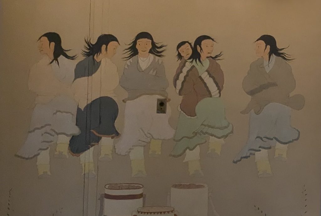 A painted mural of four Apache women dancing with baskets in the foreground. One of the women is carrying a child on her back.