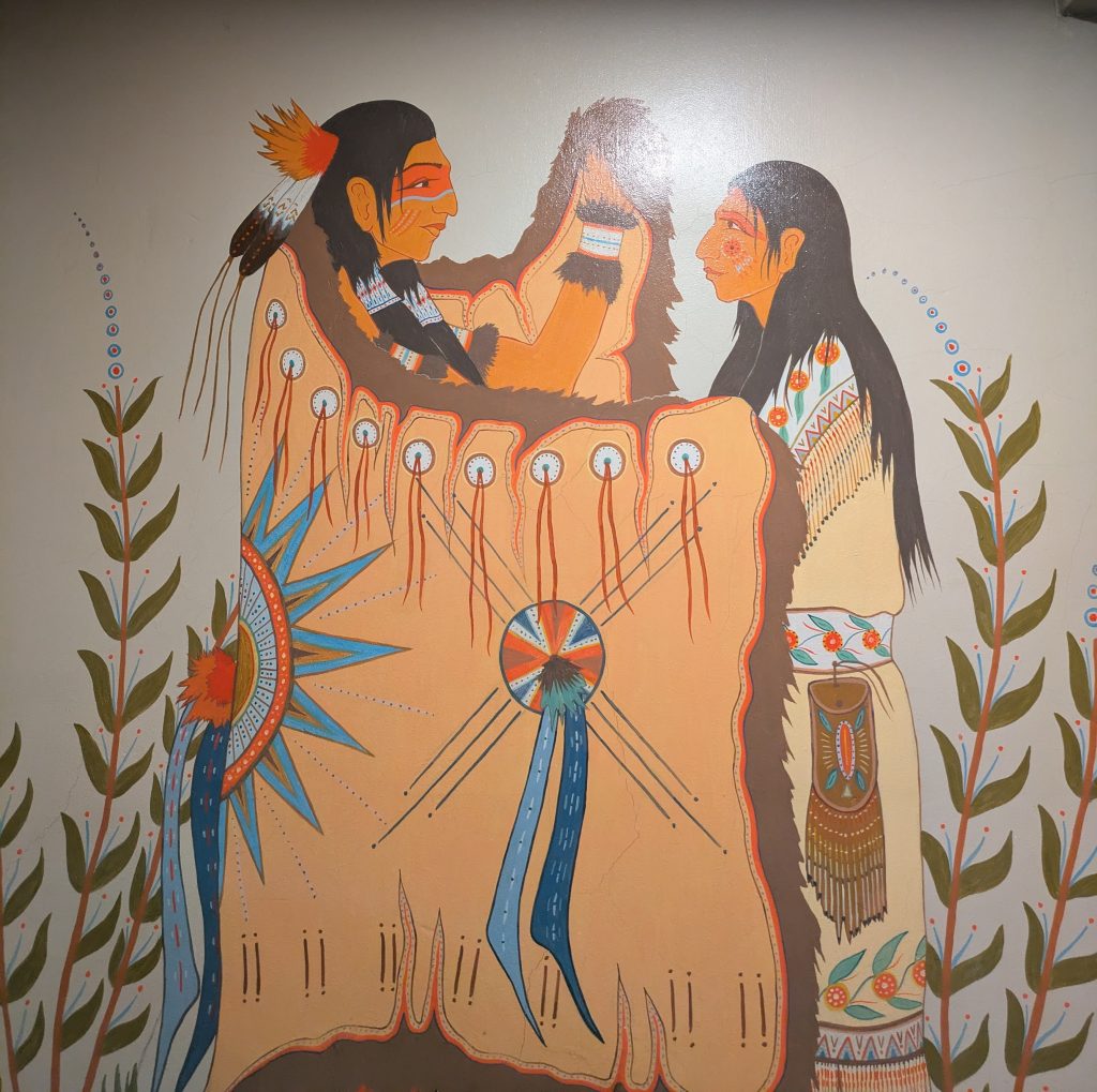 A painted mural of a Potawotamie man in traditional regalia wrapping a Potawotamie woman in traditional regalia in an animal hide.