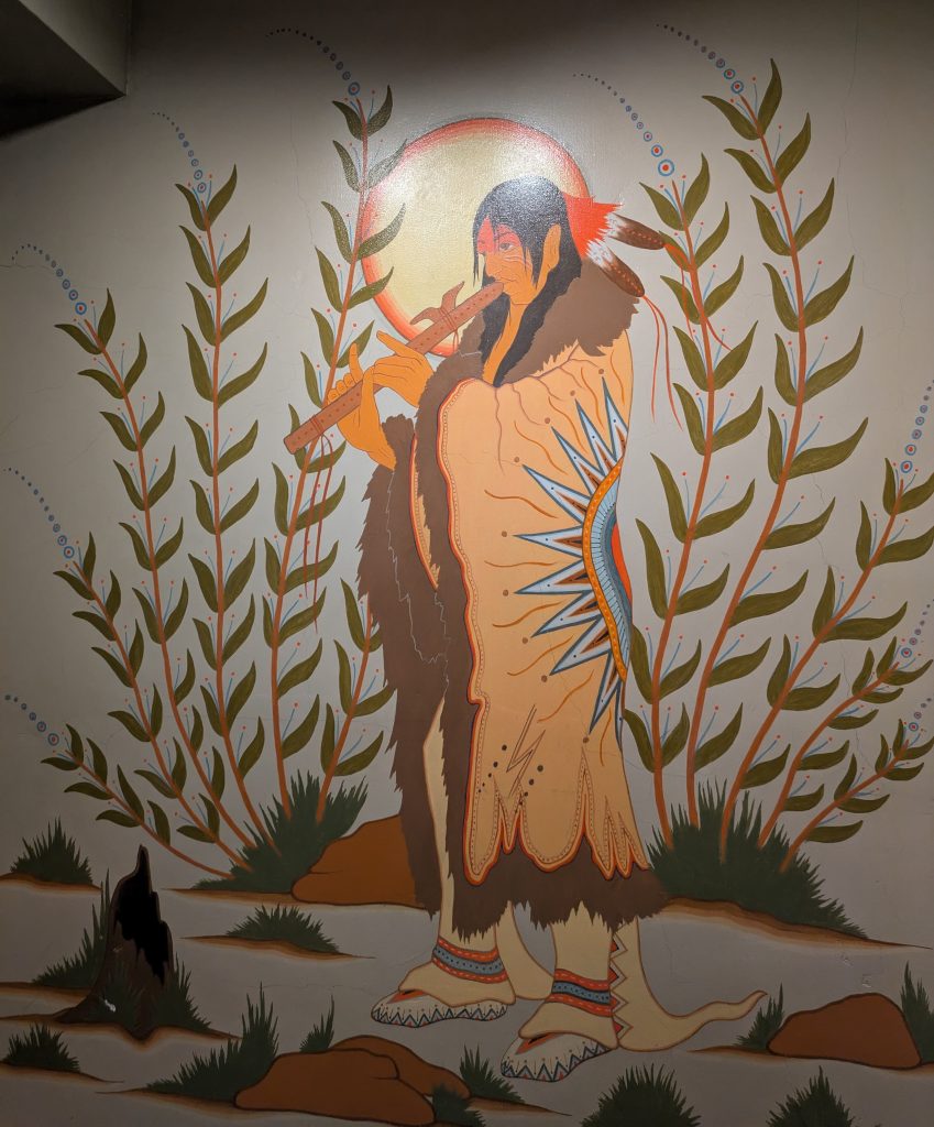 A Potawotamie man wearing traditional regalia playing a flute surrounded by plants with the sun in the background.